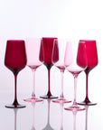 Six pink and magenta tinted wine glasses arranged in a row on a reflective white surface against a white background.