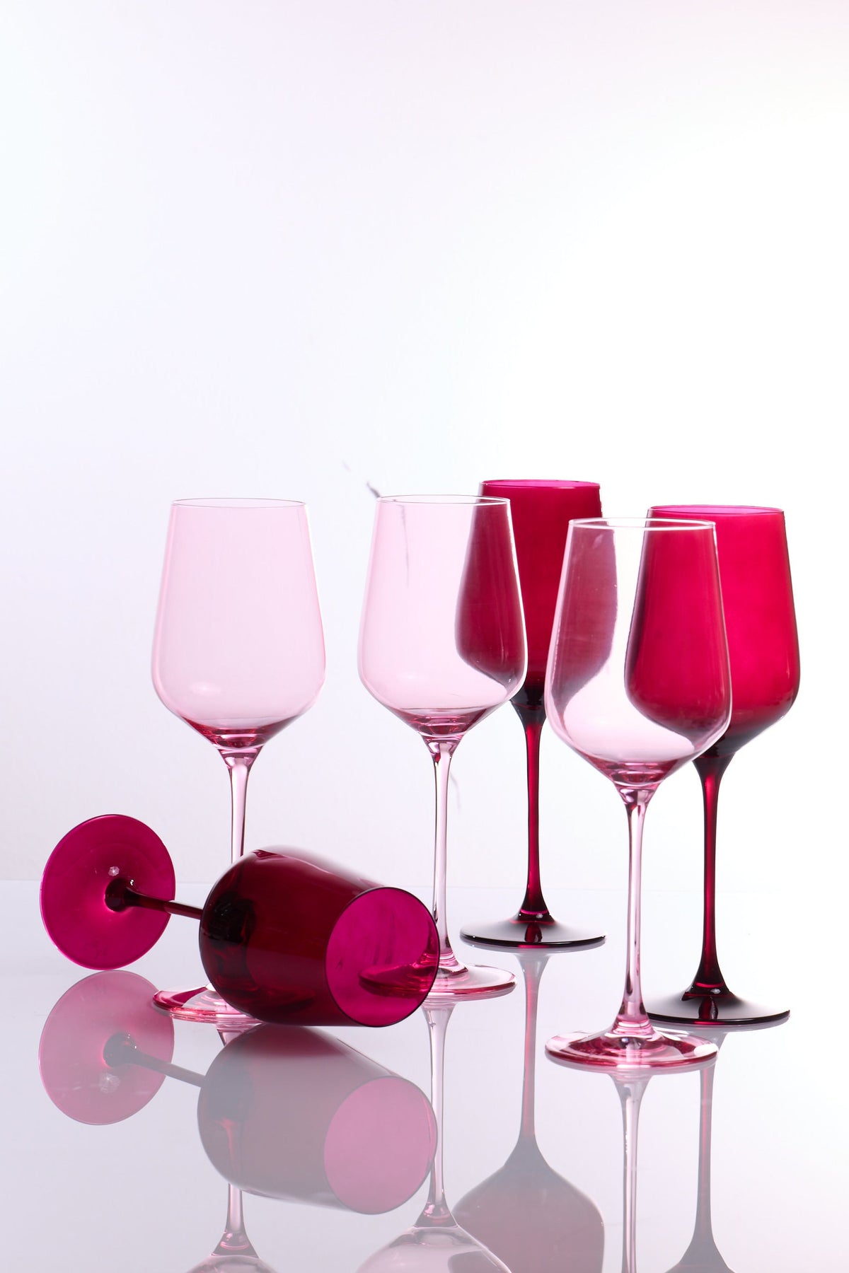 Six pink and magenta tinted wine glasses arranged on a glossy white reflective surface against a white studio background.