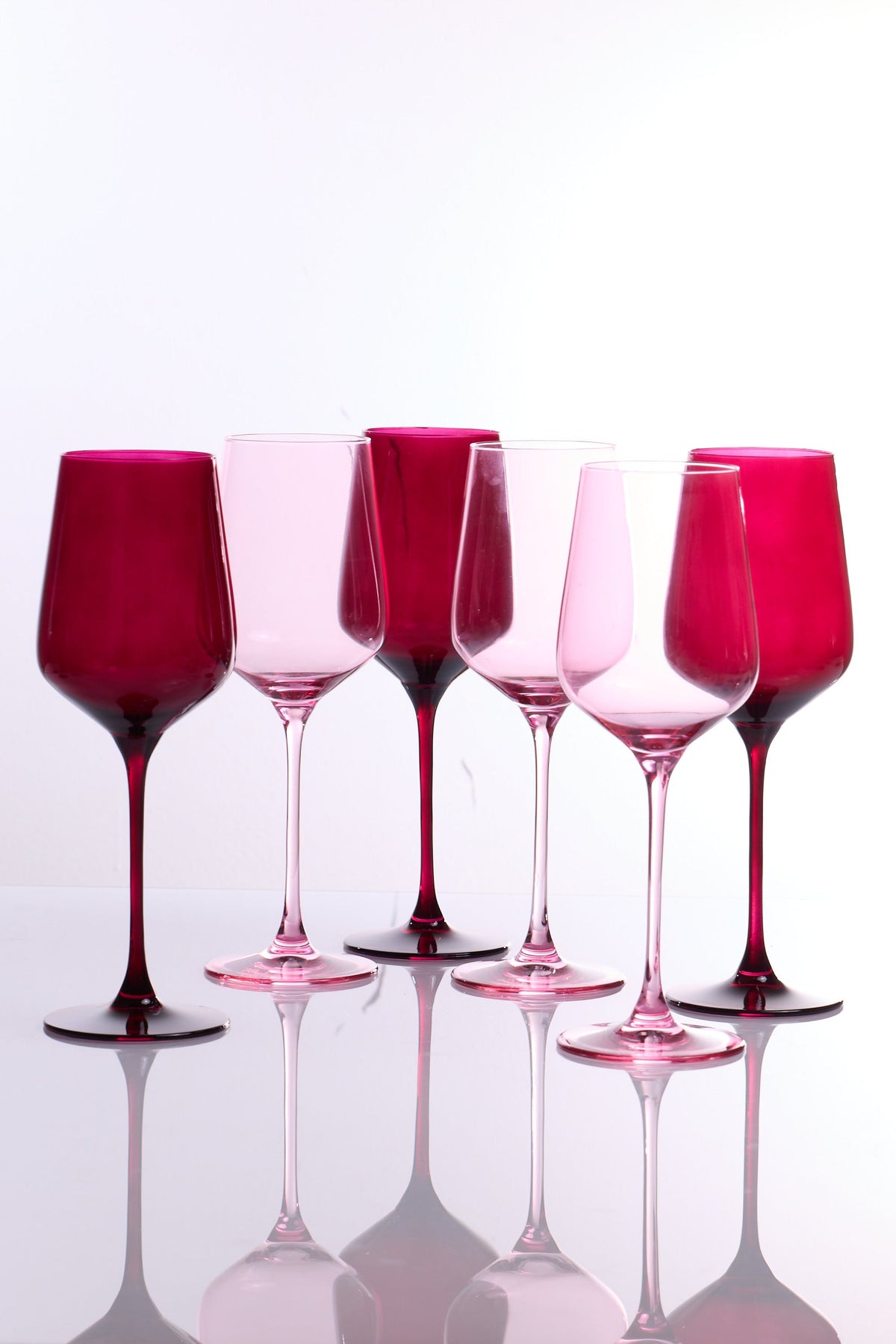 Six pink and magenta tinted wine glasses arranged in a row on a reflective white surface against a white background.