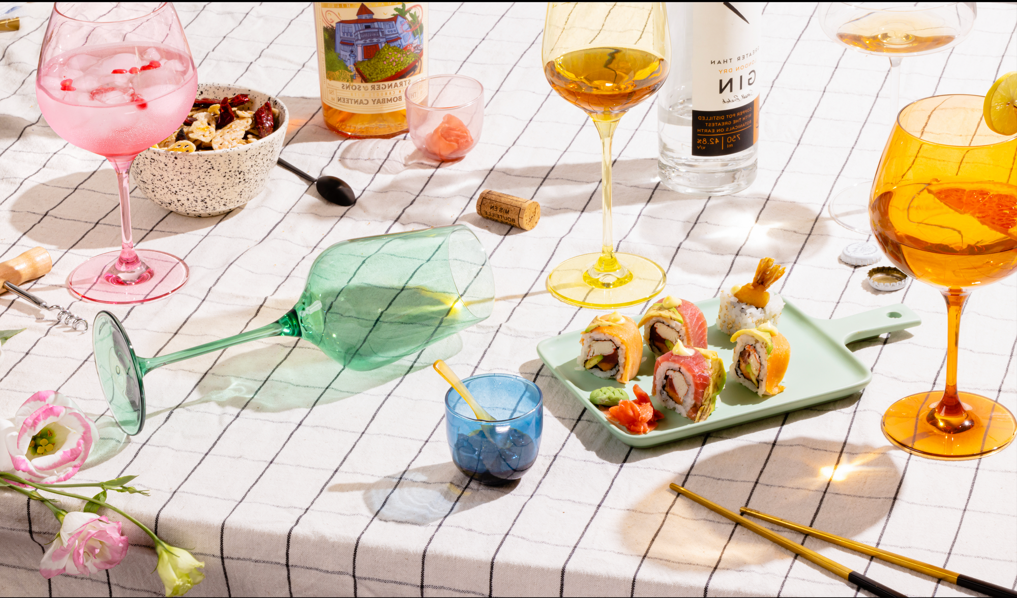 Table setting with drinks, sushi, and flowers on a checkered tablecloth.